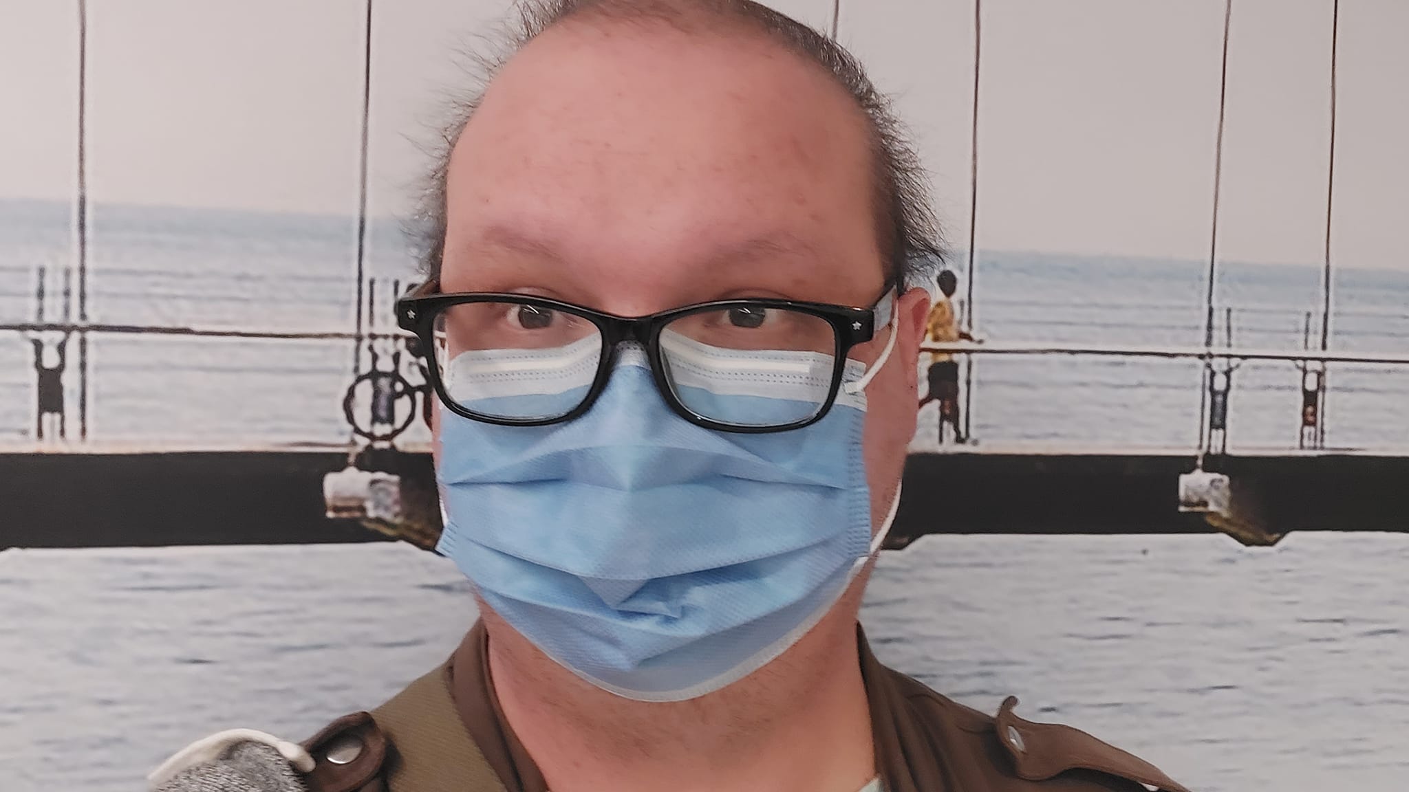 A man wearing a sweater and surgical mask sits in front of a suspension bridge poster in a hospital waiting room